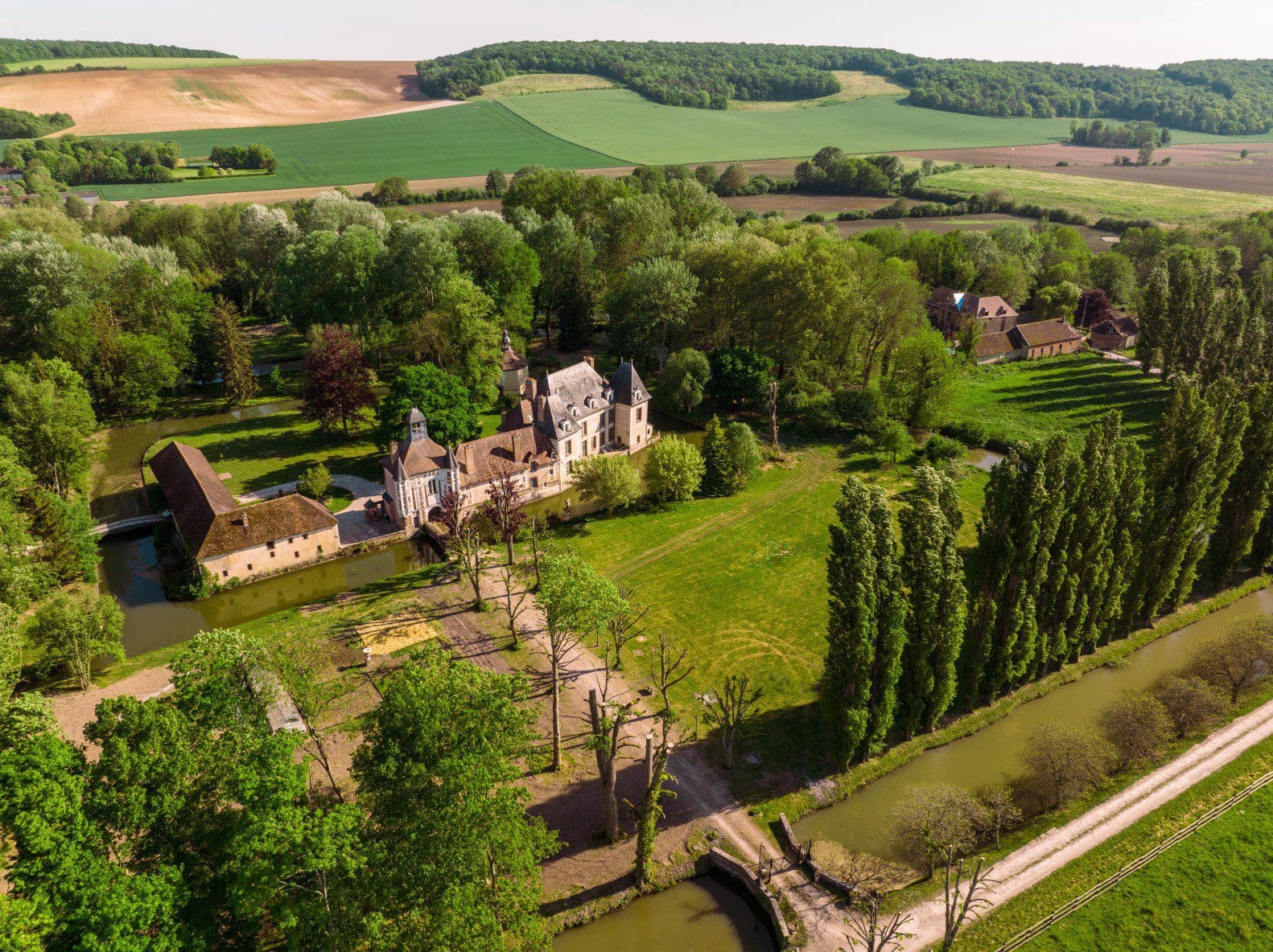 UN DOUBLE RÉSEAU DE DOUVES.<br />
Sur la rivière Vanne et le bief qui alimente le moulin, un premier réseau de douves très larges isole le château et ses communs campés dans un terreplein dont le pigeonnier se détache presque. Un second réseau plus vaste enserre le verger et le po tager bordé au nord et à l’ouest par un spectaculaire alignement de peupliers d’Italie, sans doute plantés par l’industriel et homme politique troyen Charles Buxtorf et son épouse, Mlle d’Ambly, héritière de Saint-Benoist.<br />
Studio OG